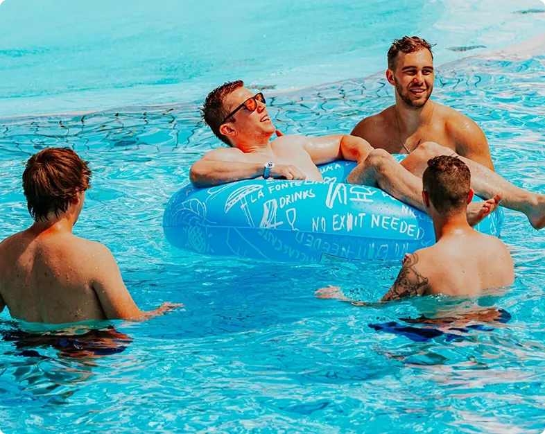 Vier junge Männer entspannen im Pool, einer liegt auf einem blauen Schwimmring mit weißem Schriftzug.