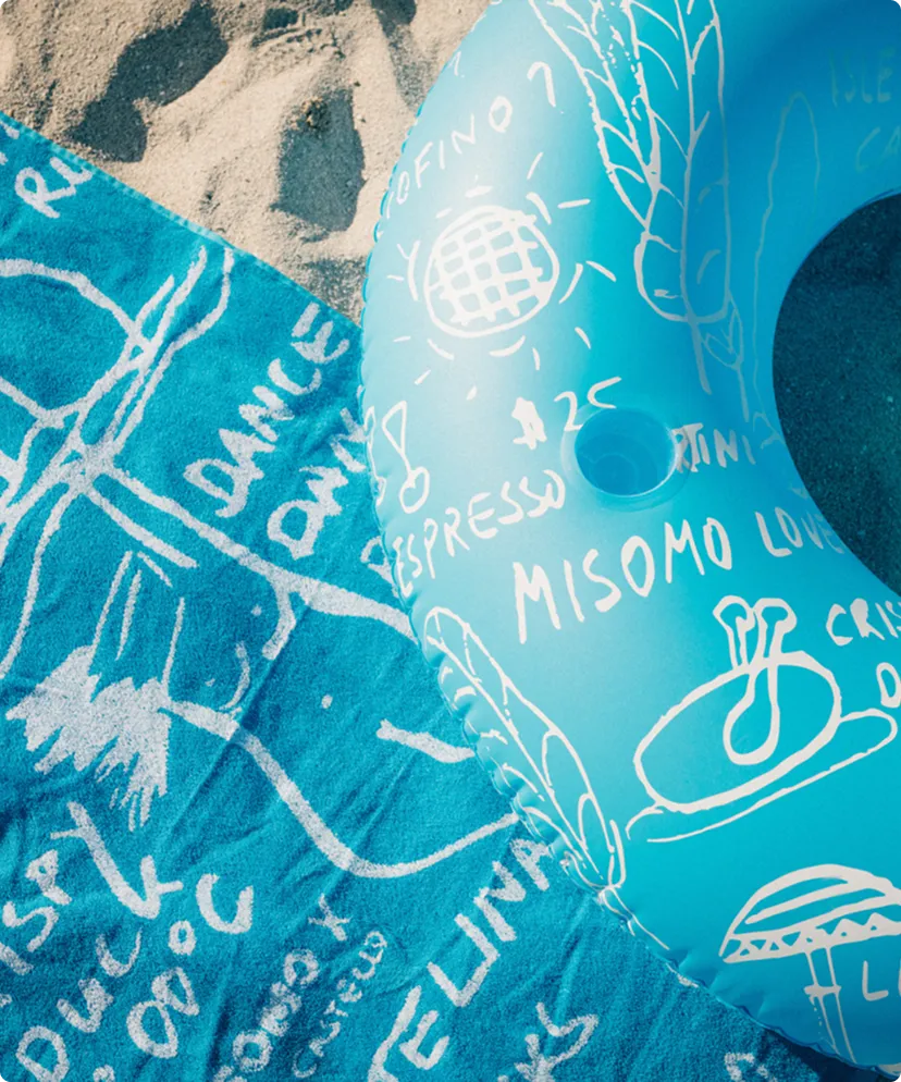 Blauer aufblasbarer Schwimmring mit weißen Kritzelzeichnungen und Texten liegt auf einem blauen Handtuch mit weißen Zeichnungen auf Sand.