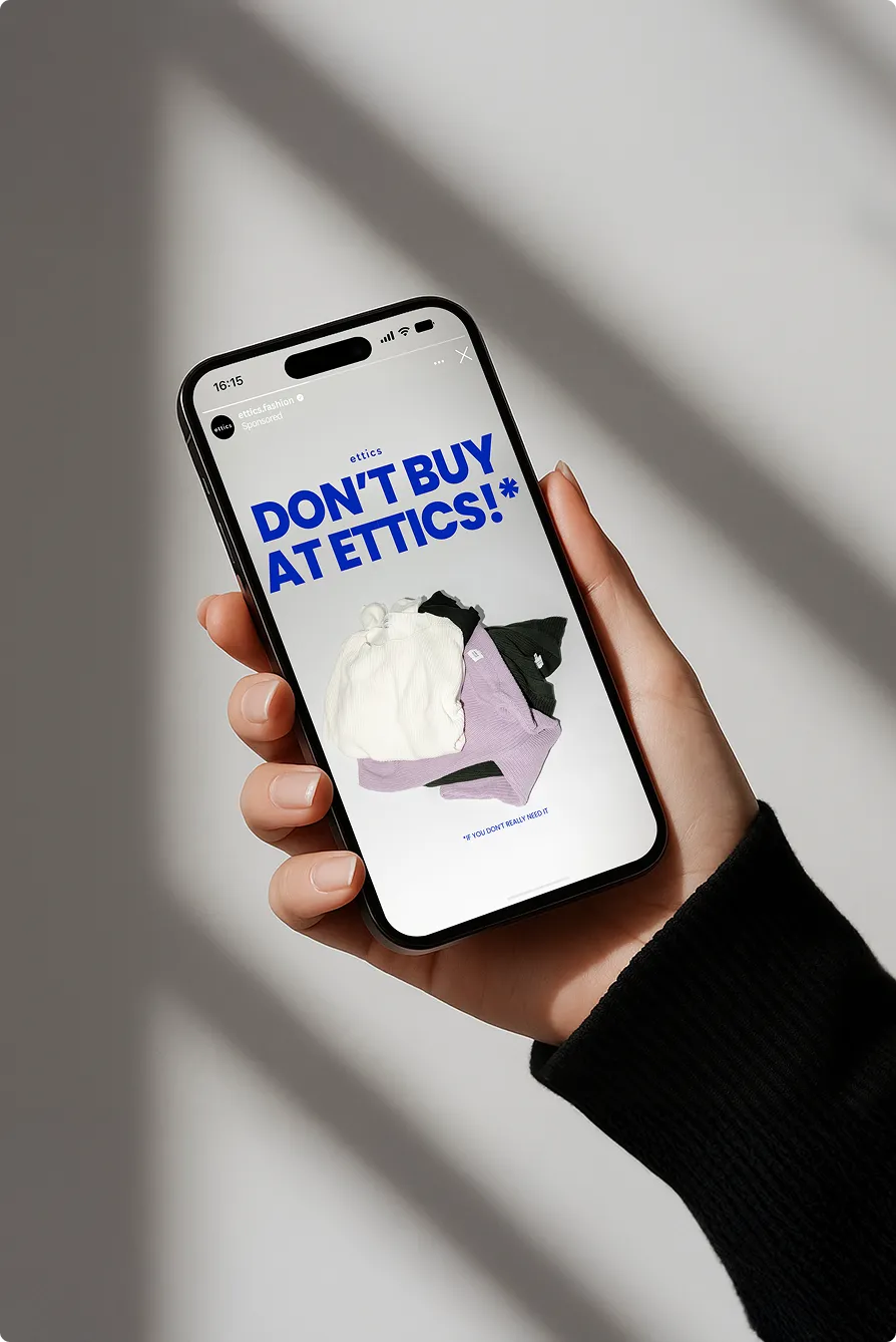 Hand hält ein Smartphone mit einer Anzeige von ettics, die in blauer Schrift sagt: "Kaufe nicht bei ettics!", darunter liegen einige gefaltete Pullover.