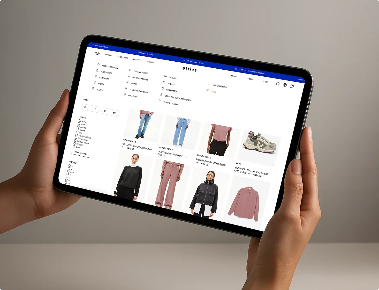 Person hält ein Tablet mit einer Online-Shop-Seite für nachhaltige Kleidung, die Kleidungskategorien und Produktbilder zeigt.