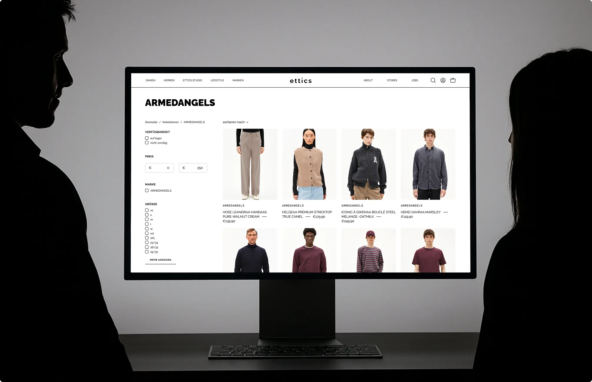 Zwei Personen betrachten einen Desktop-Computerbildschirm, der eine Online-Shop-Seite für ARMEDANGELS-Kleidung zeigt.