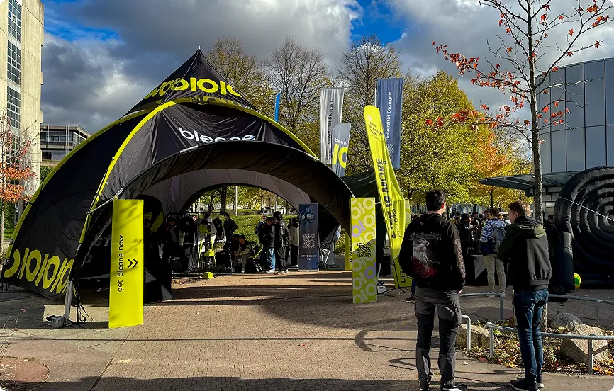 Menschen unter und um ein großes schwarzes und gelbes Zelt mit Aufschrift 'bleeno' auf einem Platz mit Bäumen und Gebäuden bei bewölktem Himmel.