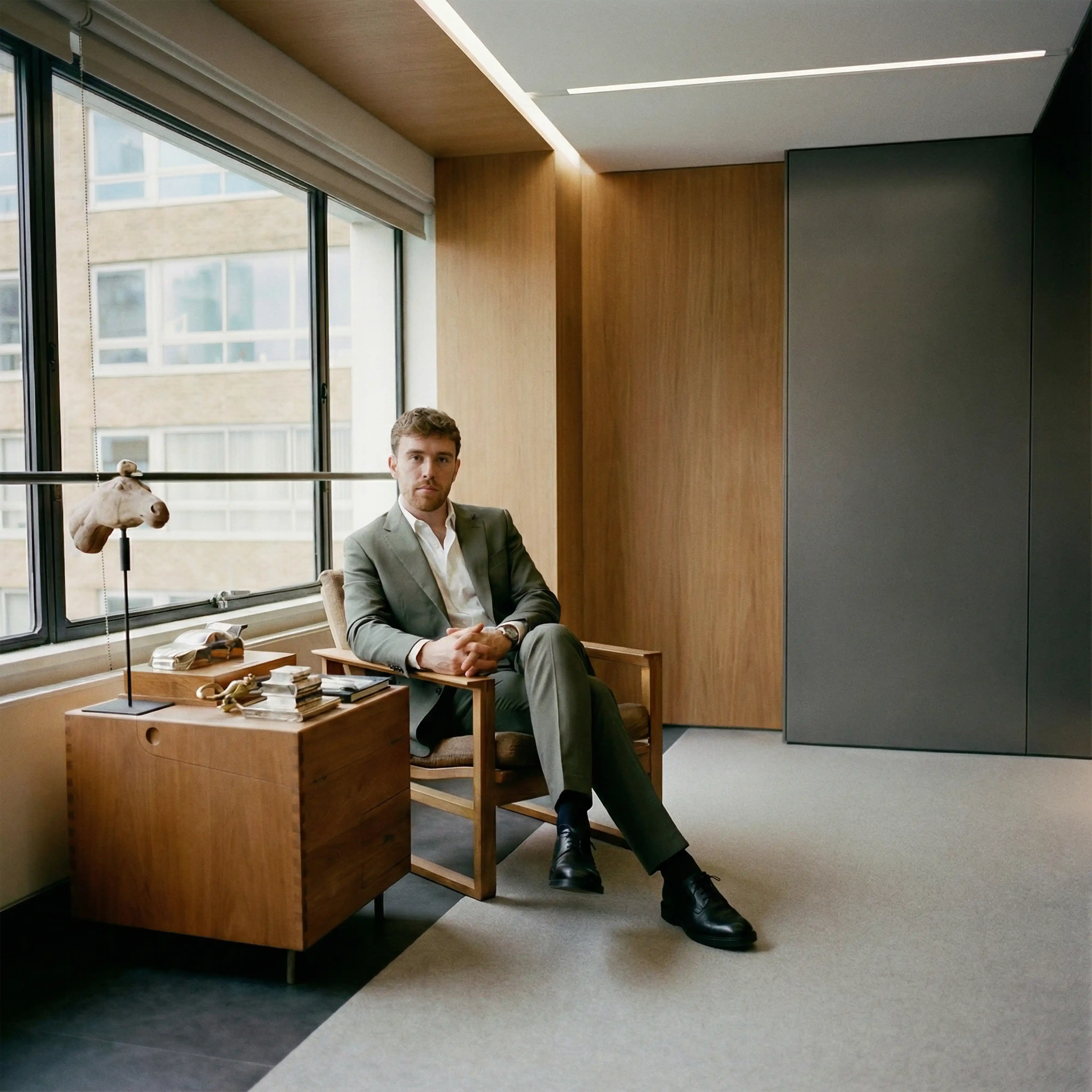 Ein Mann im grauen Anzug sitzt auf einem Holzstuhl in einem modernen Raum mit großen Fenstern und einem Holztisch neben ihm.