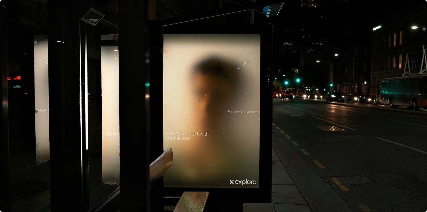 Eine unscharfe Werbetafel an einer Bushaltestelle bei Nacht zeigt die Silhouette eines Gesichts und den Text „Clarity can start with a small step“ sowie „Premium Self Drug-Testing“ und das Logo von Exploro.