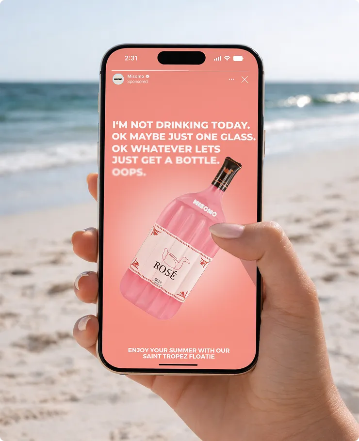 Hand hält ein Smartphone mit Werbung eines rosafarbenen Weinflaschen-Designs am Strand.