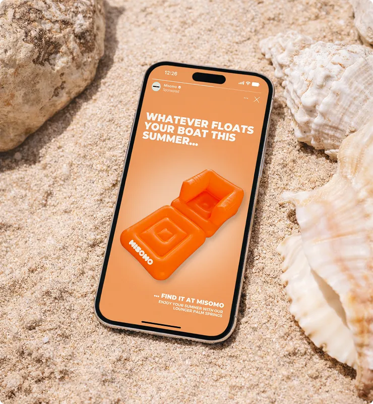 Smartphone liegt im Sand zwischen Muscheln, zeigt Werbung für orangefarbene aufblasbare Liege mit Text 'Whatever floats your boat this summer' und Hinweis auf Misomo.