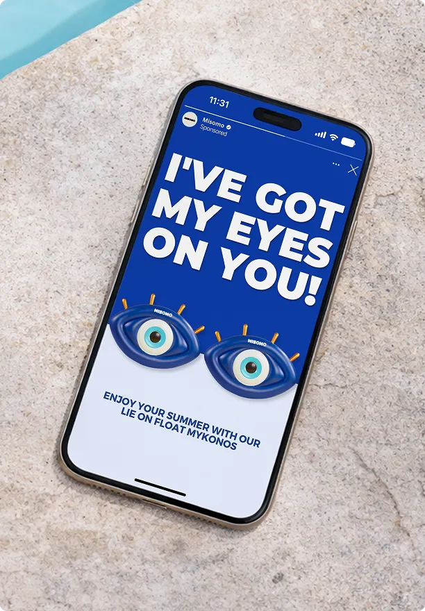 Ein Smartphone zeigt eine Anzeige mit dem Text 'I'VE GOT MY EYES ON YOU!' und zwei blauen aufblasbaren Augen mit der Aufschrift 'MISOMO' darunter, die Sommerwerbung für 'Lie on Float Mykonos' darstellt.