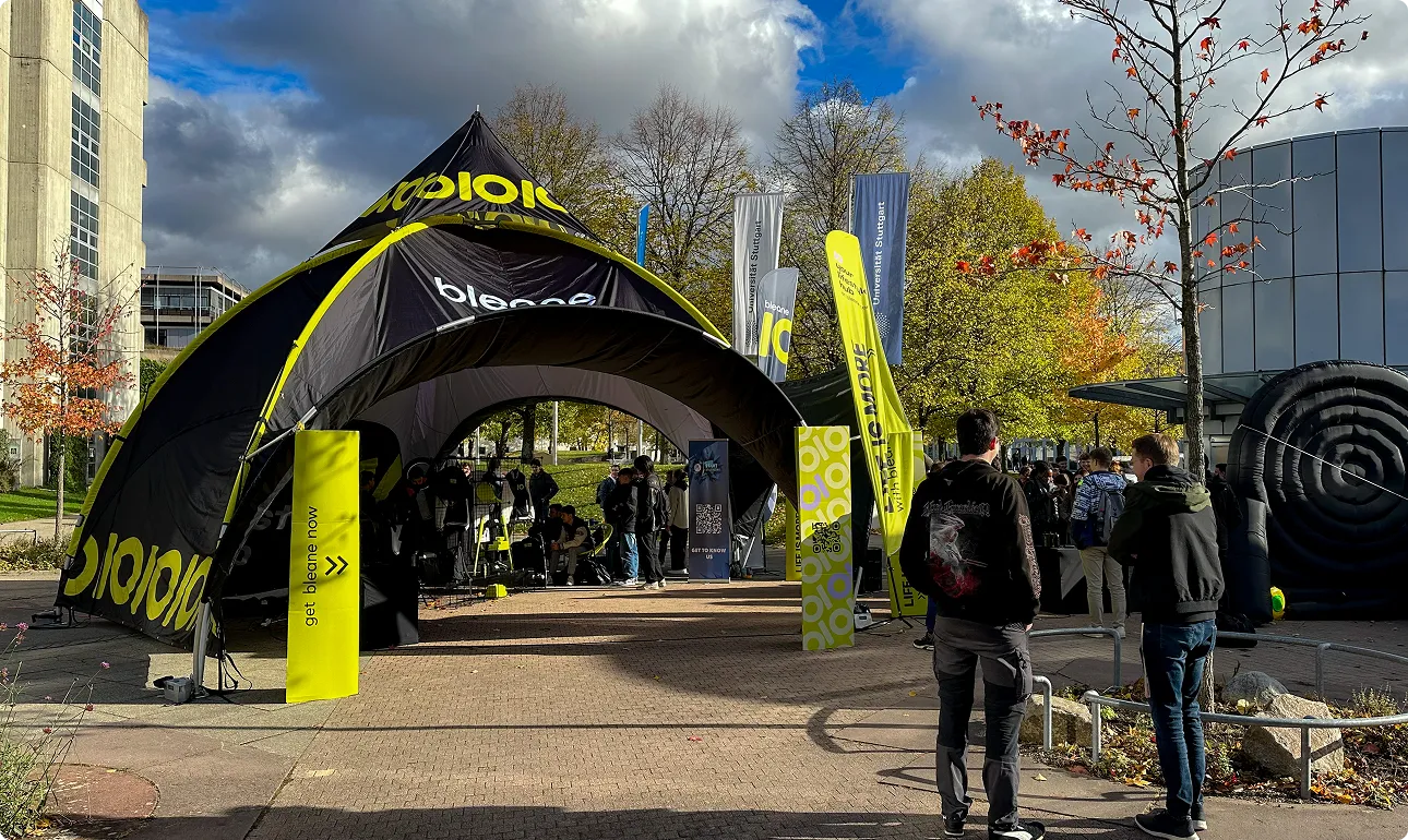Schwarzes und gelbes Zelt mit Aufschrift 'bleone' auf einem Campus, umgeben von Menschen und herbstlich gefärbten Bäumen.