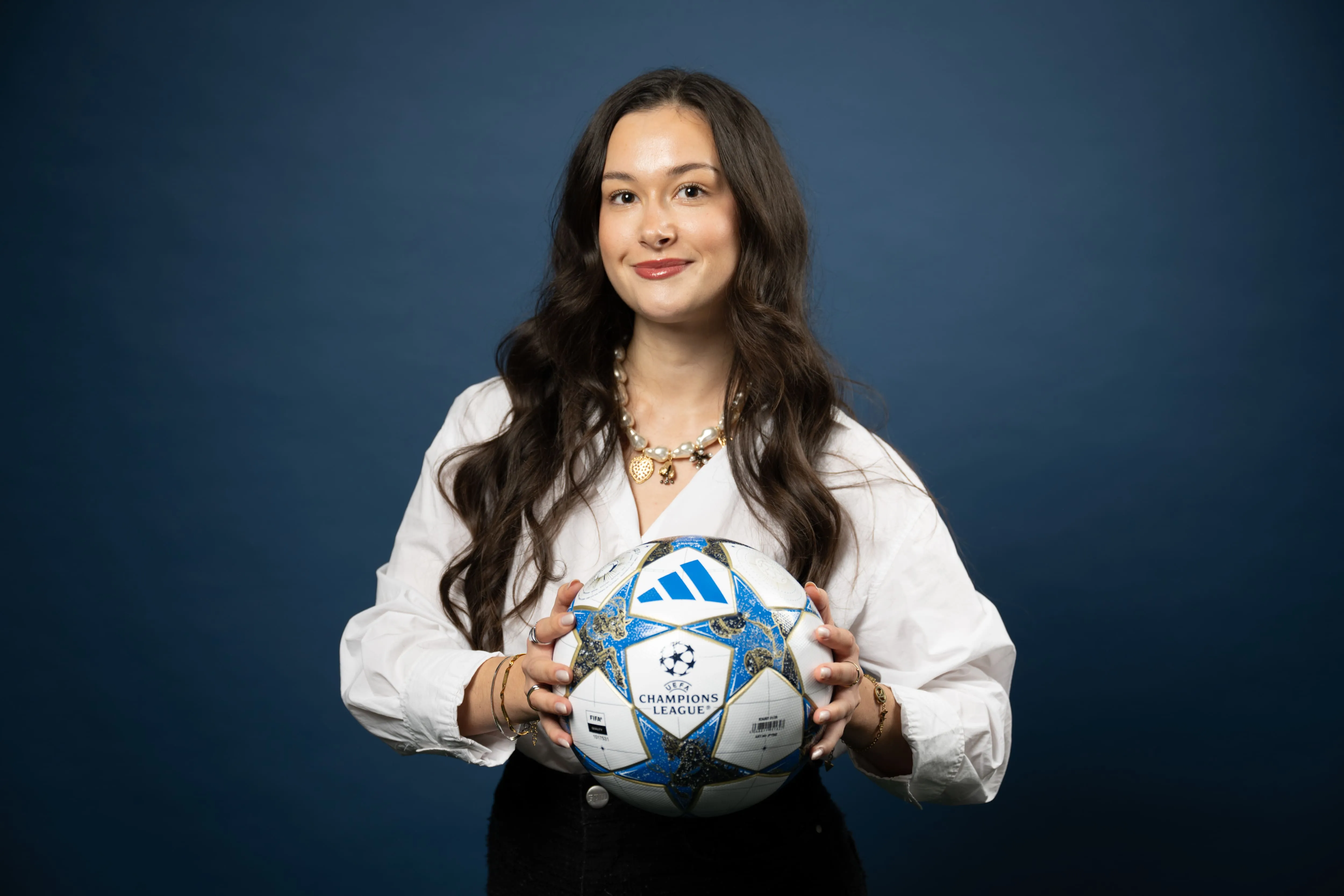 Junge Frau mit langen braunen Haaren hält einen UEFA Champions League Fußball vor einem dunklen Hintergrund.