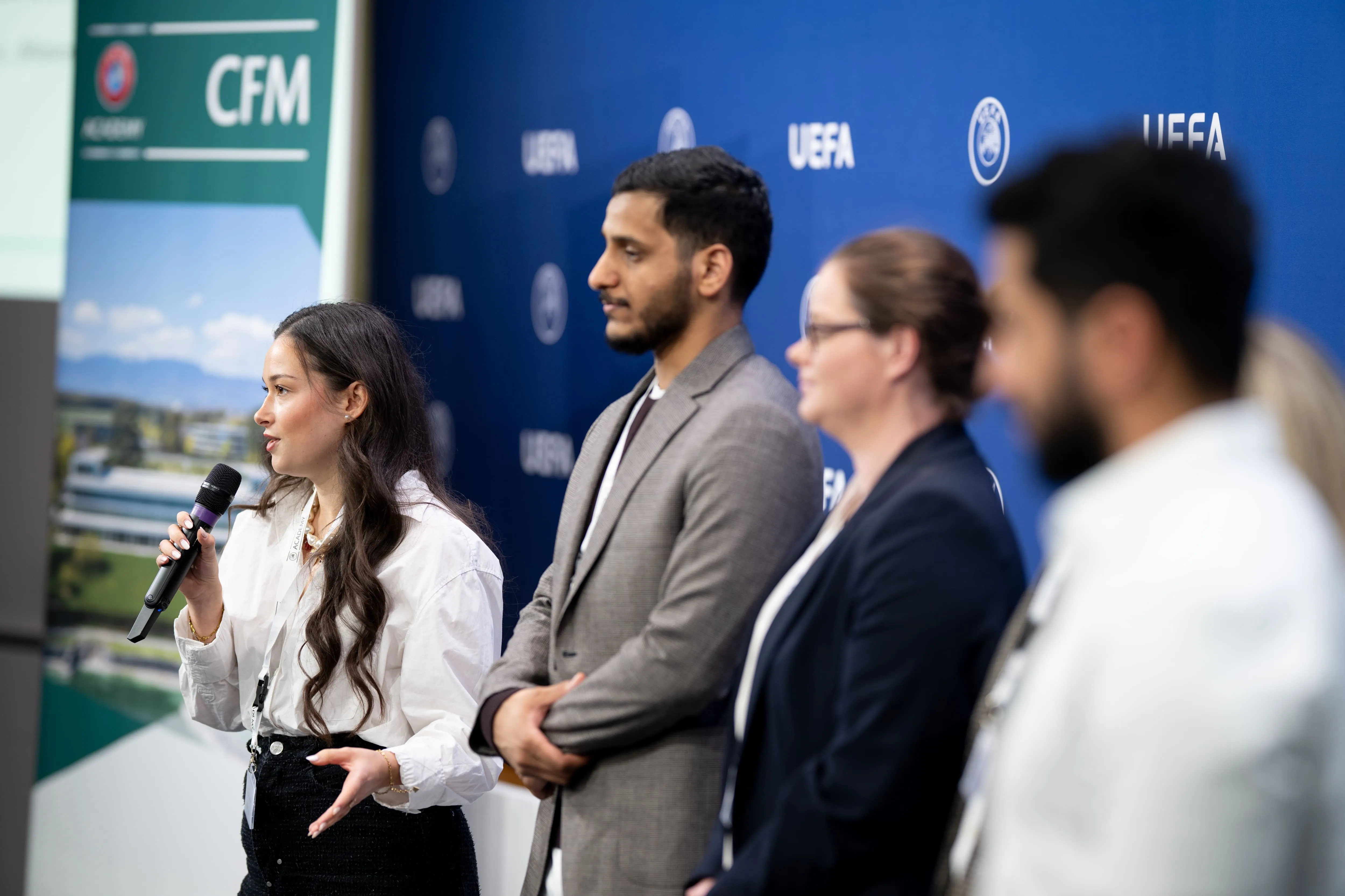 Frau spricht mit Mikrofon vor drei stehenden Personen vor einer blauen UEFA-Hintergrundwand.