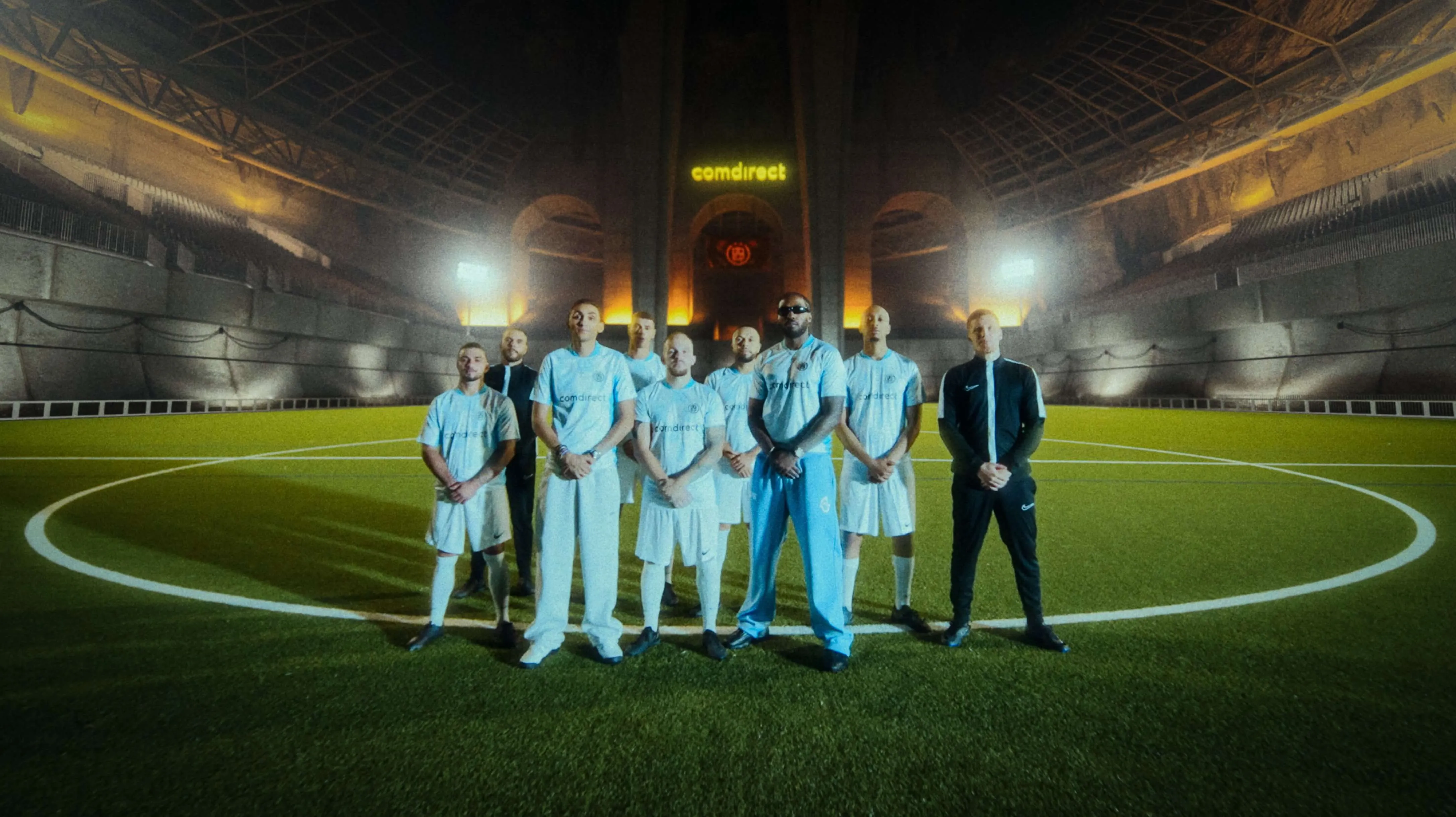 Ein Fußballteam steht in weißen Trikots und Trainingsanzügen auf einem beleuchteten Spielfeld in einer großen Halle mit dem Schriftzug comdirect im Hintergrund.