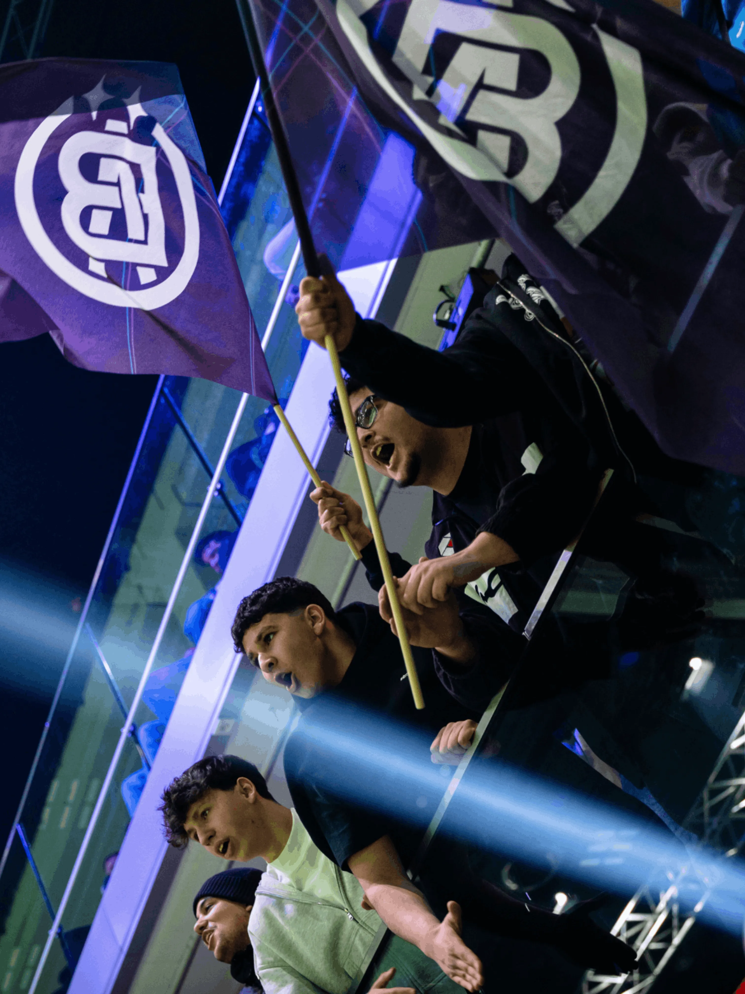 Vier junge Männer jubeln enthusiastisch bei einem Event, zwei halten Fahnen mit weißem Logo vor dunklem Hintergrund.