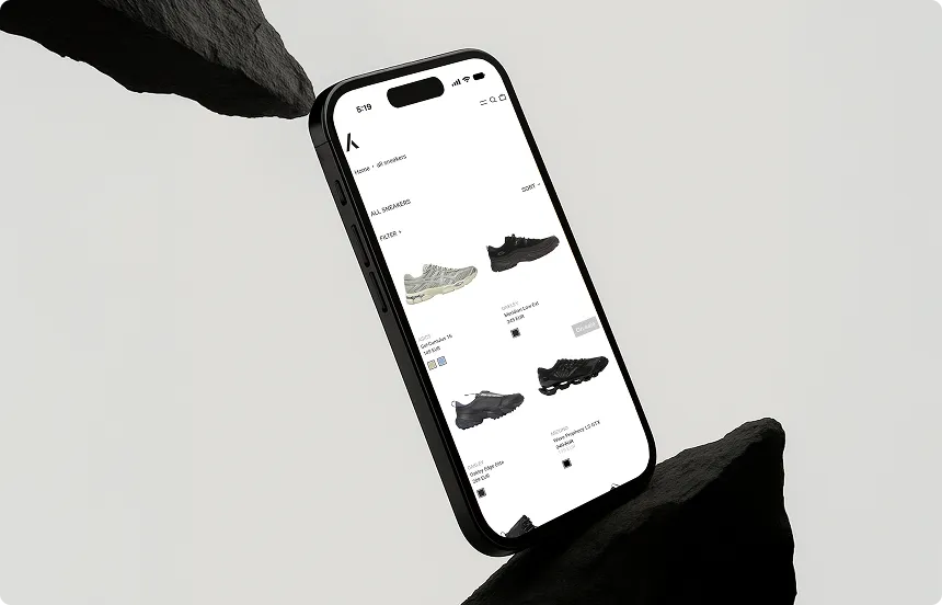 Smartphone zeigt eine Webseite mit verschiedenen Sneakers in einer minimalistischen, schwarz-grauen Umgebung zwischen zwei schwarzen Felsen.