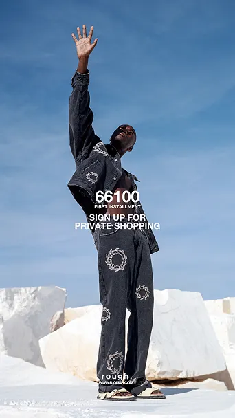 Person im schwarzen Outfit mit Muster hebt eine Hand gegen den blauen Himmel, vor großen weißen Felsen.