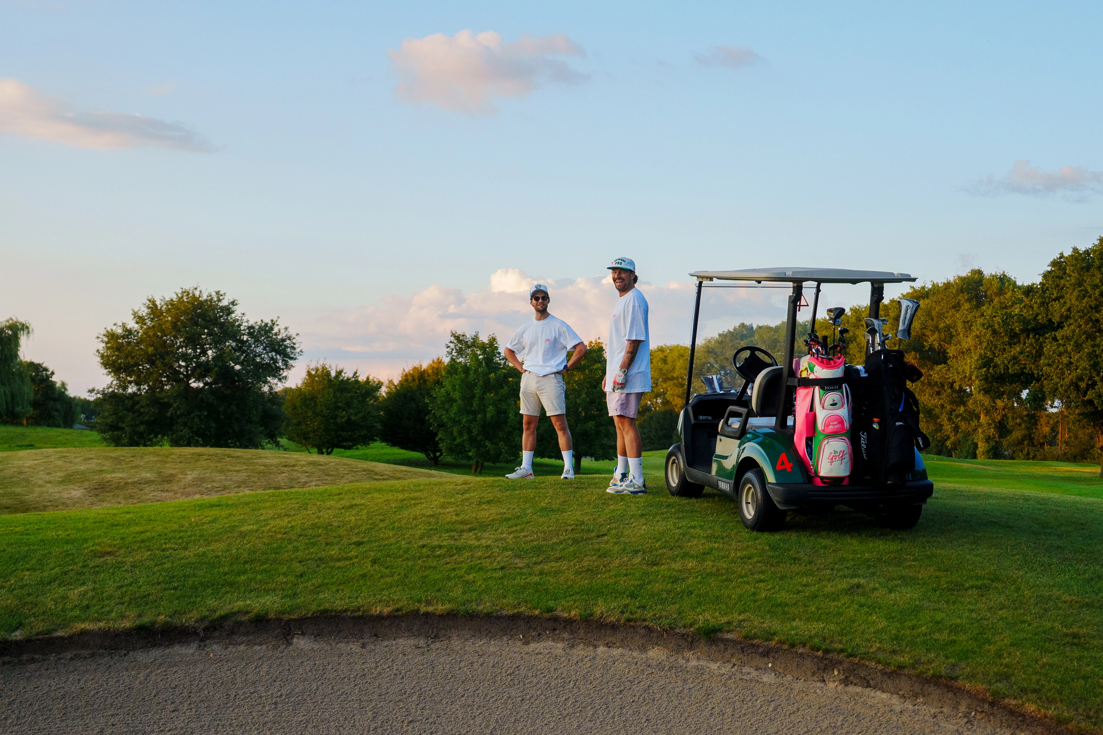 Zwei Männer in Sommerkleidung stehen neben einem Golfwagen mit Golfschlägern auf einem grünen Golfplatz.