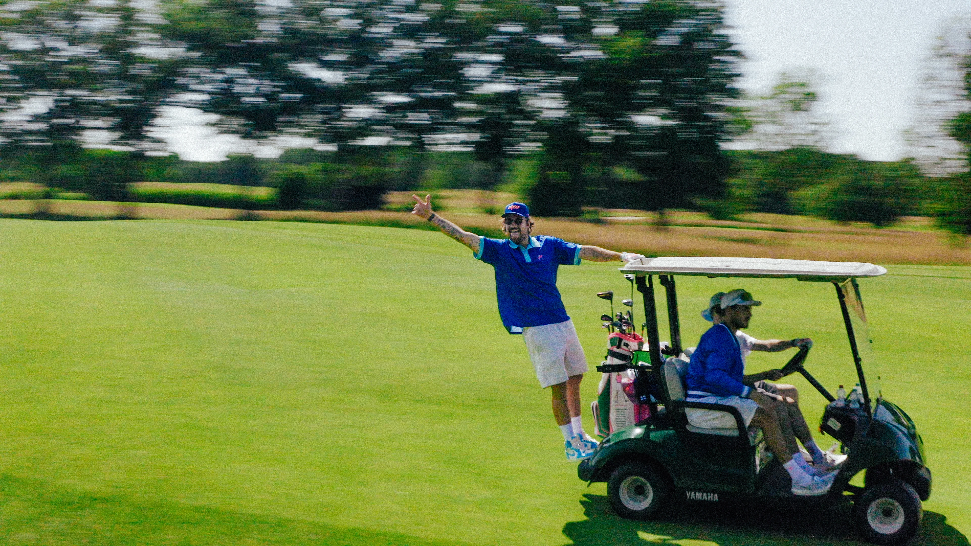 Drei Männer auf einem Golfwagen fahren über einen Rasen, einer steht hinten am Wagen und winkt fröhlich.