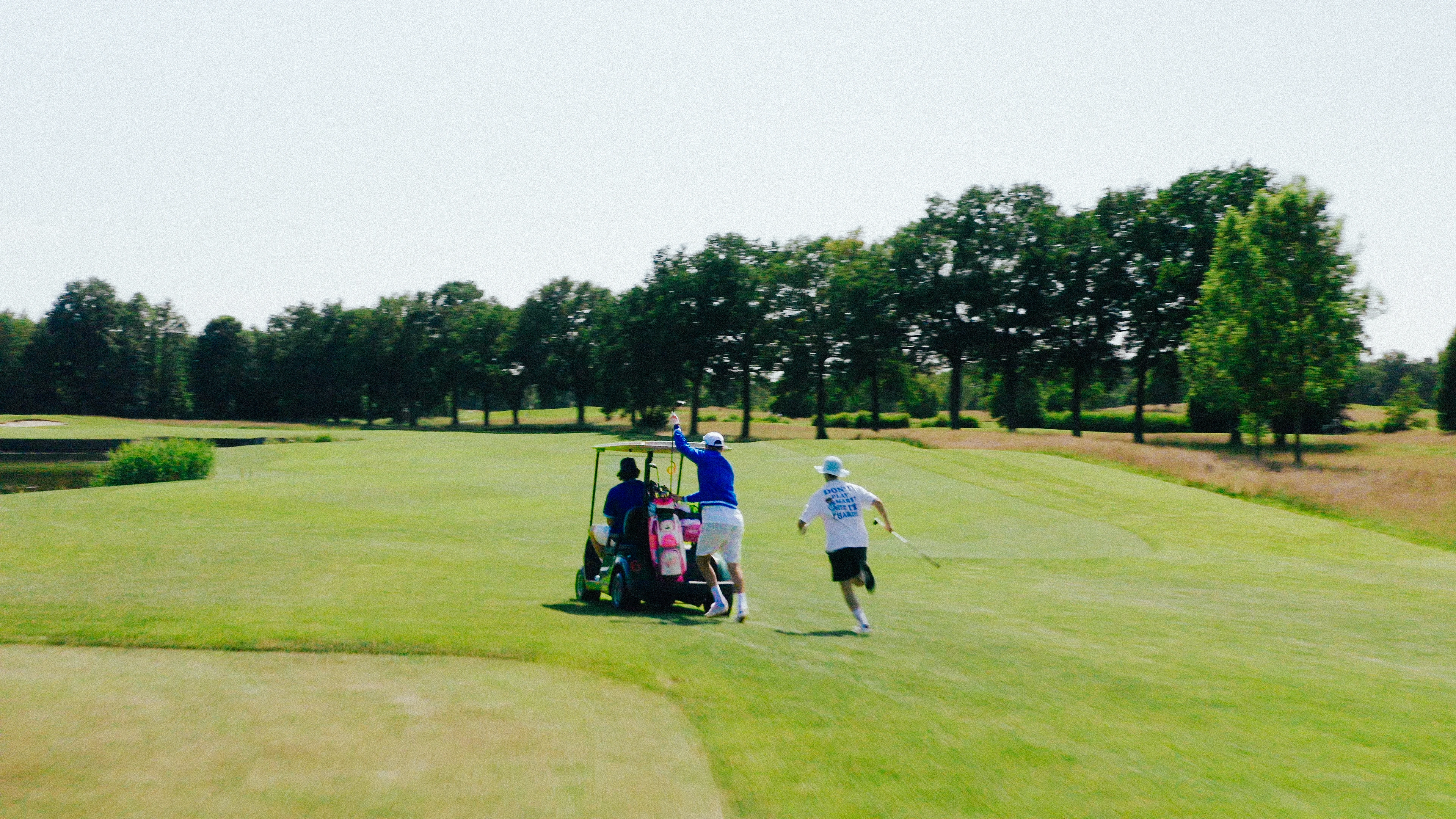 Drei Golfer auf einem Golfplatz, zwei rennen neben einem Golfwagen mit Schlägern, umgeben von Bäumen.