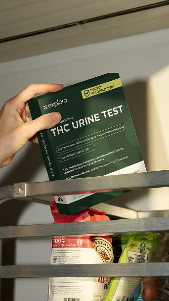 Hand hält eine grüne Explor Testbox für THC-Urin mit der Aufschrift "Doctor Recommended" vor einem Kühlschrank.