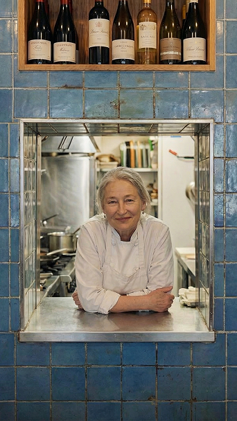 Ältere Köchin mit weißen Haaren und weißer Schürze lehnt lächelnd am Durchreichefenster in einer Küche mit blauen Fliesen und Weinflaschenregal darüber.