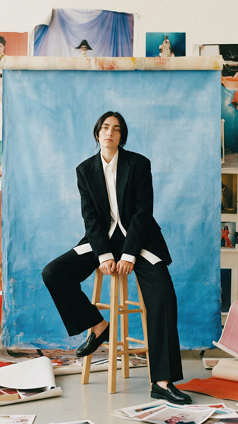 Person mit schwarzen kurzen Haaren in schwarzem Anzug und weißen Hemd sitzt auf einem Holzstuhl vor blauem Hintergrund, umgeben von verstreuten Papierbögen und Fotodrucken.