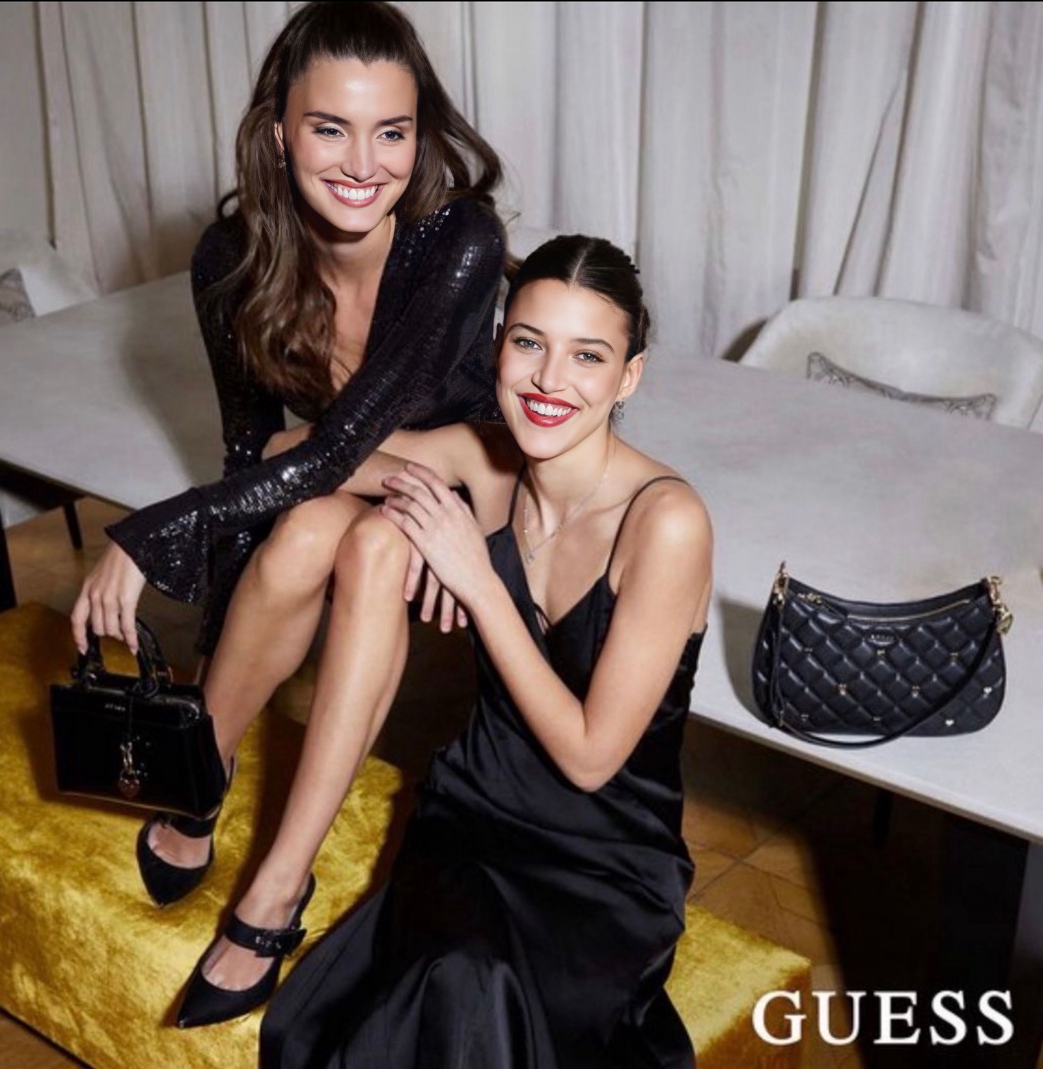 Zwei lächelnde Frauen in eleganten schwarzen Kleidern sitzen auf einem gelben Sitzhocker, eine hält eine schwarze Handtasche, eine weitere schwarze Tasche liegt auf einem Tisch im Hintergrund.