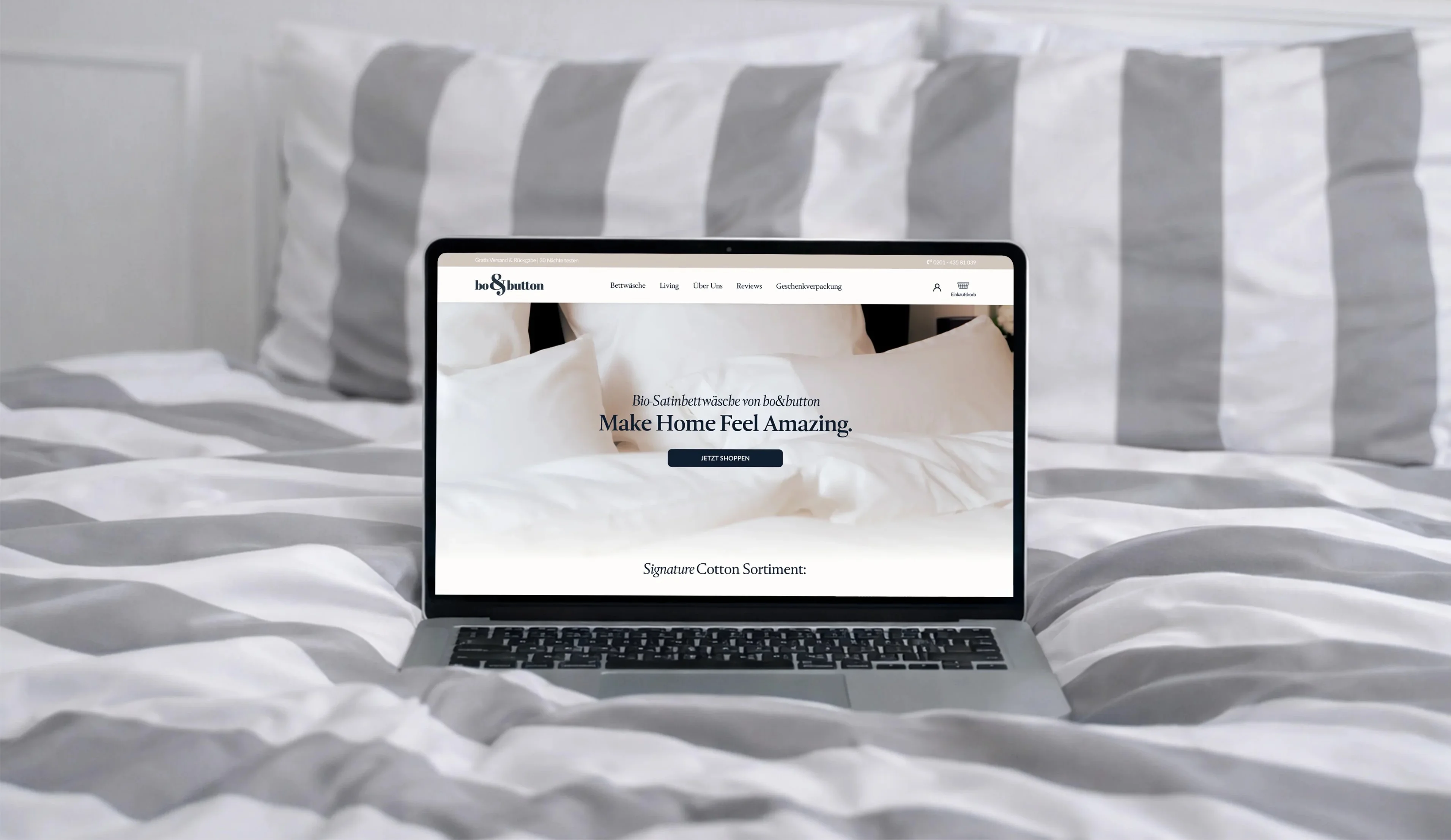 Laptop zeigt bo&button Website für Bio-Satinbettwäsche auf gestreifter Bettdecke in Weiß und Grau.