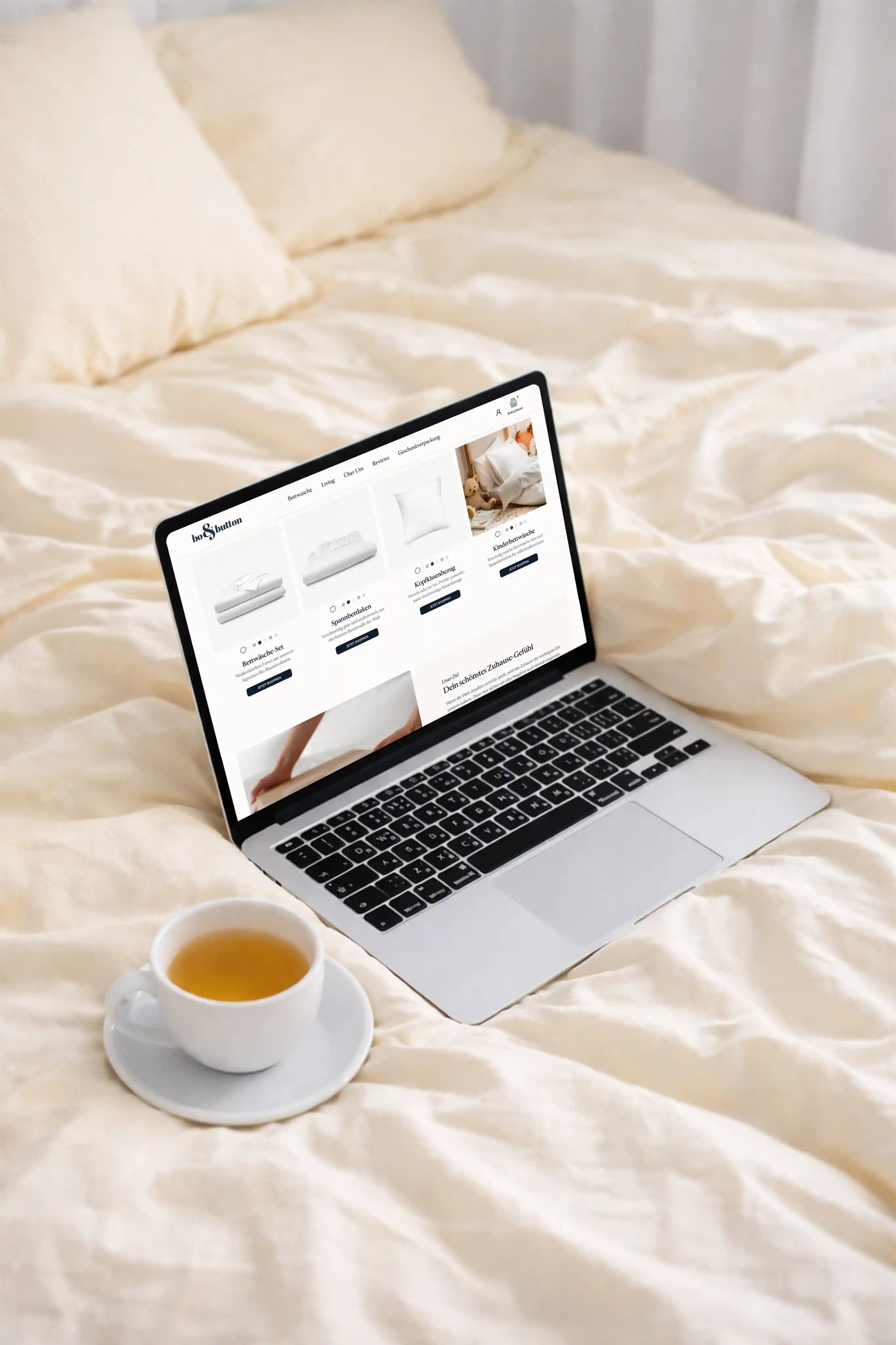 Laptop auf einem cremefarbenen Bett mit einer Website für Bettwaren und eine Tasse Tee auf einem Unterteller daneben.
