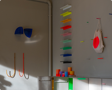 Farbiges modernes Wandregal mit Haken, Taschen und bunten Rollen an einer hellen Wand im Sonnenlicht.