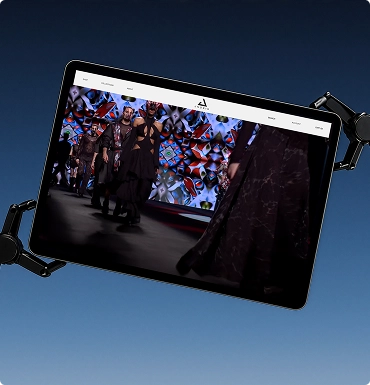 Tablet held by robotic arms displaying a runway fashion show with models walking in dark and patterned outfits.