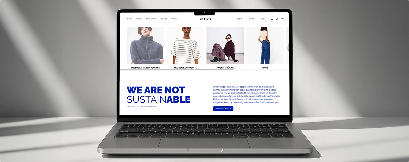 Laptop zeigt eine nachhaltige Mode-Website mit Kategorien Pullover, Kleider, Hosen und Jeans sowie dem Text ‚We are not sustainable‘.