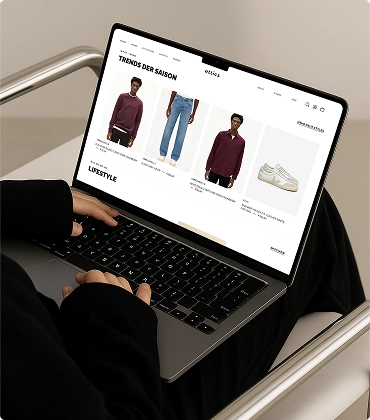 Person scrolling on a laptop showing a fashion website with men's clothing and shoes categorized under 'Trends der Saison'.