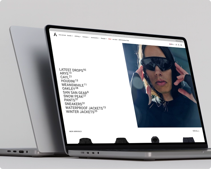 Laptop mit geöffneter Webseite eines Outdoor-Bekleidungsshops, zeigt Kategorien wie neueste Produkte, Sneaker und Jacken sowie ein Bild einer Person mit Sonnenbrille und Kapuzenpullover.