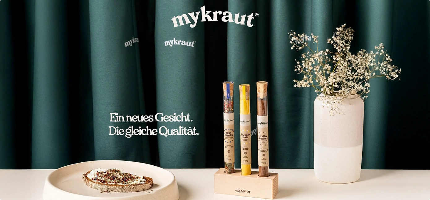 Drei mykraut Gewürzröhrchen in einem Holzständer neben einer Scheibe Brot mit Aufstrich und einer weißen Vase mit Trockenblumen vor grünen Vorhängen.
