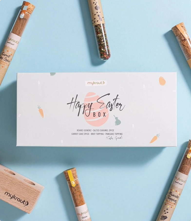 Weiße Happy Easter Box von mykraut auf hellblauem Hintergrund umgeben von Gewürzröhrchen und einem Holzhalter.