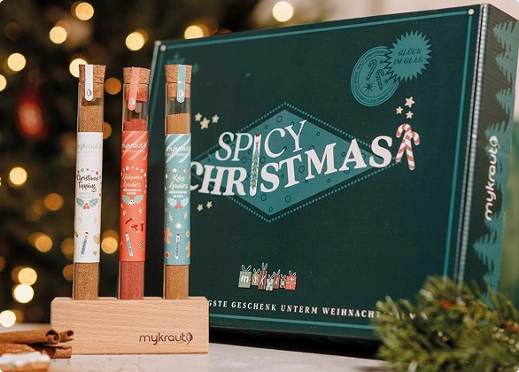 Drei Gewürzröhrchen mit Etiketten auf einem Holzständer vor einer Geschenkbox mit der Aufschrift 'Spicy Christmas'.