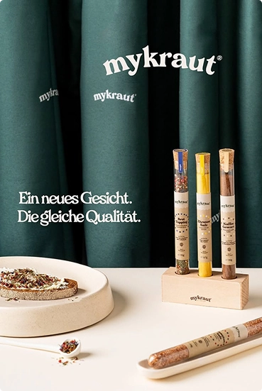 Drei Mykraut Gewürzröhrchen in einem Holzhalter vor grünem Hintergrund mit der Aufschrift Mykraut, daneben ein Teller mit Brot und einem Löffel mit Gewürzen.