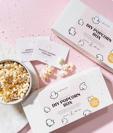 DIY Popcorn Box Verpackung mit Popcornbeutel in den Geschmacksrichtungen White Cheddar Coconut und Salted Caramel neben einer Schüssel mit frischem Popcorn auf rosafarbenem Hintergrund.