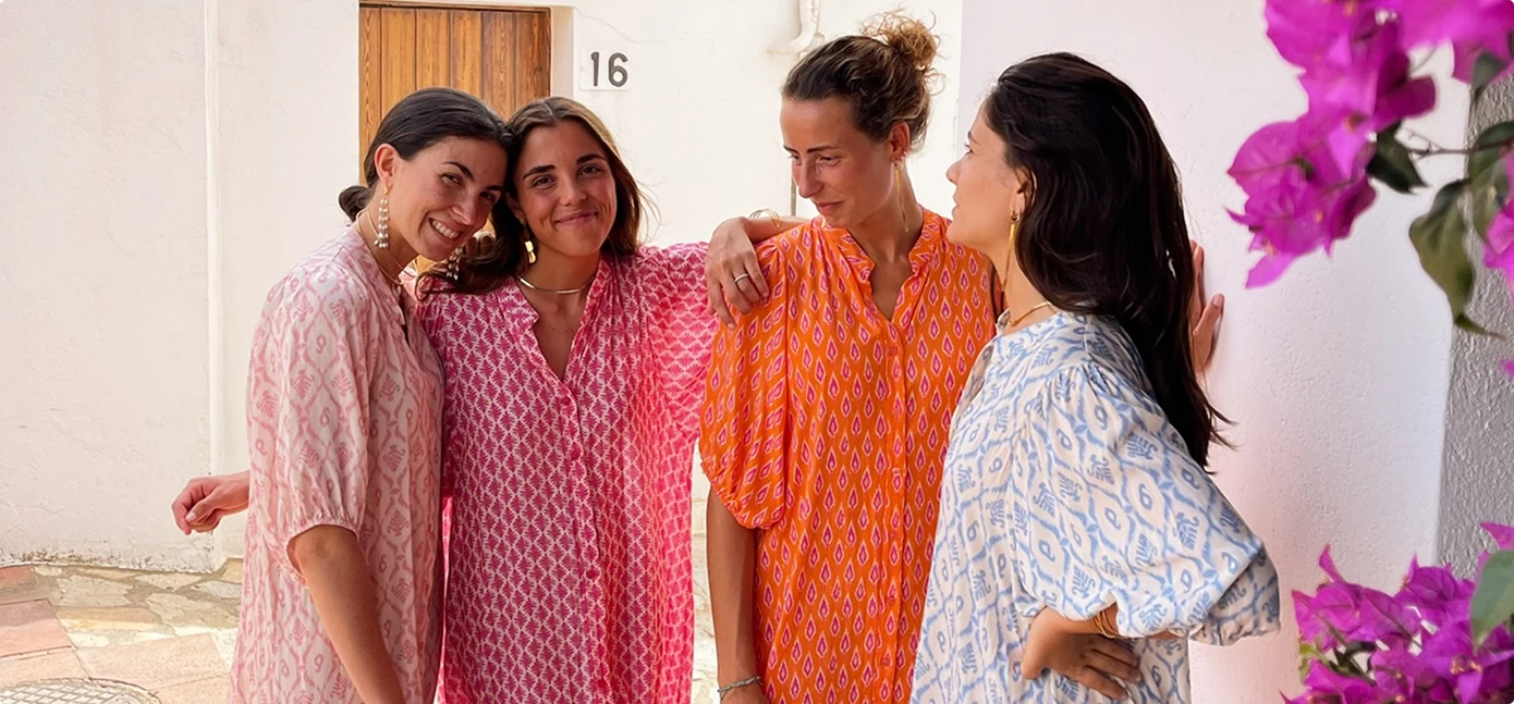 Vier Frauen in bunt gemusterten Kleidern lächeln und posieren zusammen vor einer weißen Wand mit einer Holztür und der Nummer 16, neben pinken Blüten.