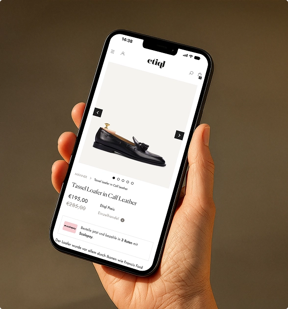 Hand hält ein Smartphone mit geöffneter Produktseite eines schwarzen Tassel Loafers aus Kalbsleder auf der Etiql-Website.