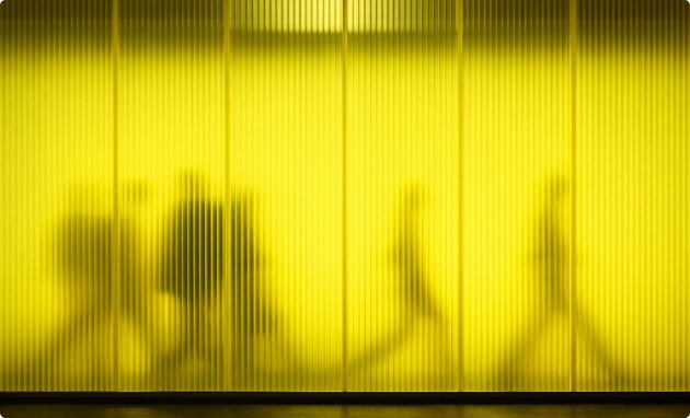 Verschwommene Schatten von Menschen, die hinter einer gelben, geriffelten Glaswand entlanggehen.