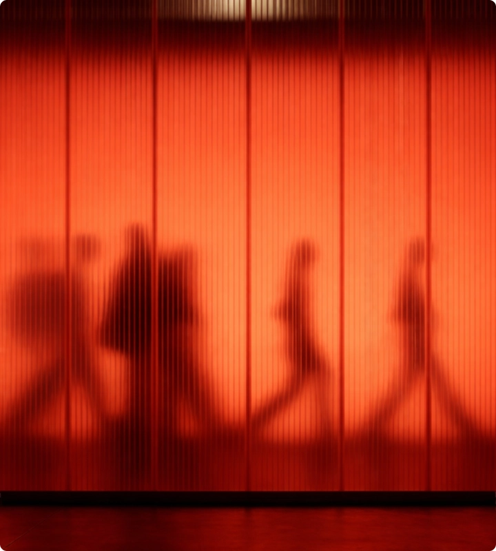 Verschwommene Silhouetten von Menschen, die vor einer roten, vertikal gestreiften Wand entlanggehen.