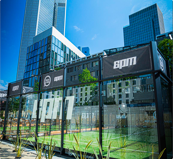 Außenansicht eines modernen Padel-Tennisplatzes mit Glaswänden und Gebäuden im Hintergrund.