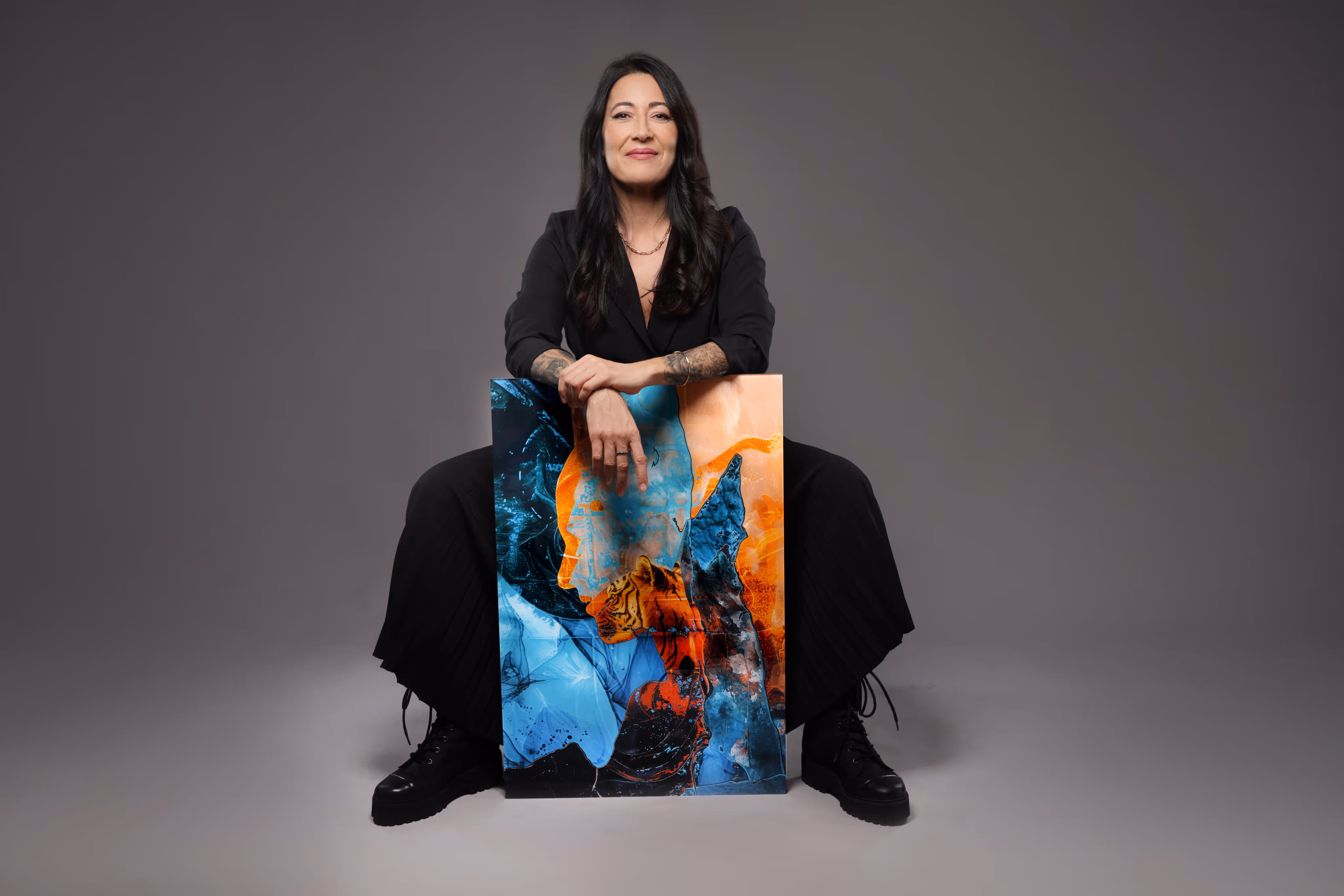 Frau mit langen dunklen Haaren in schwarzer Kleidung sitzt und hält ein farbenfrohes Kunstwerk mit blauen und orangen abstrakten Mustern.