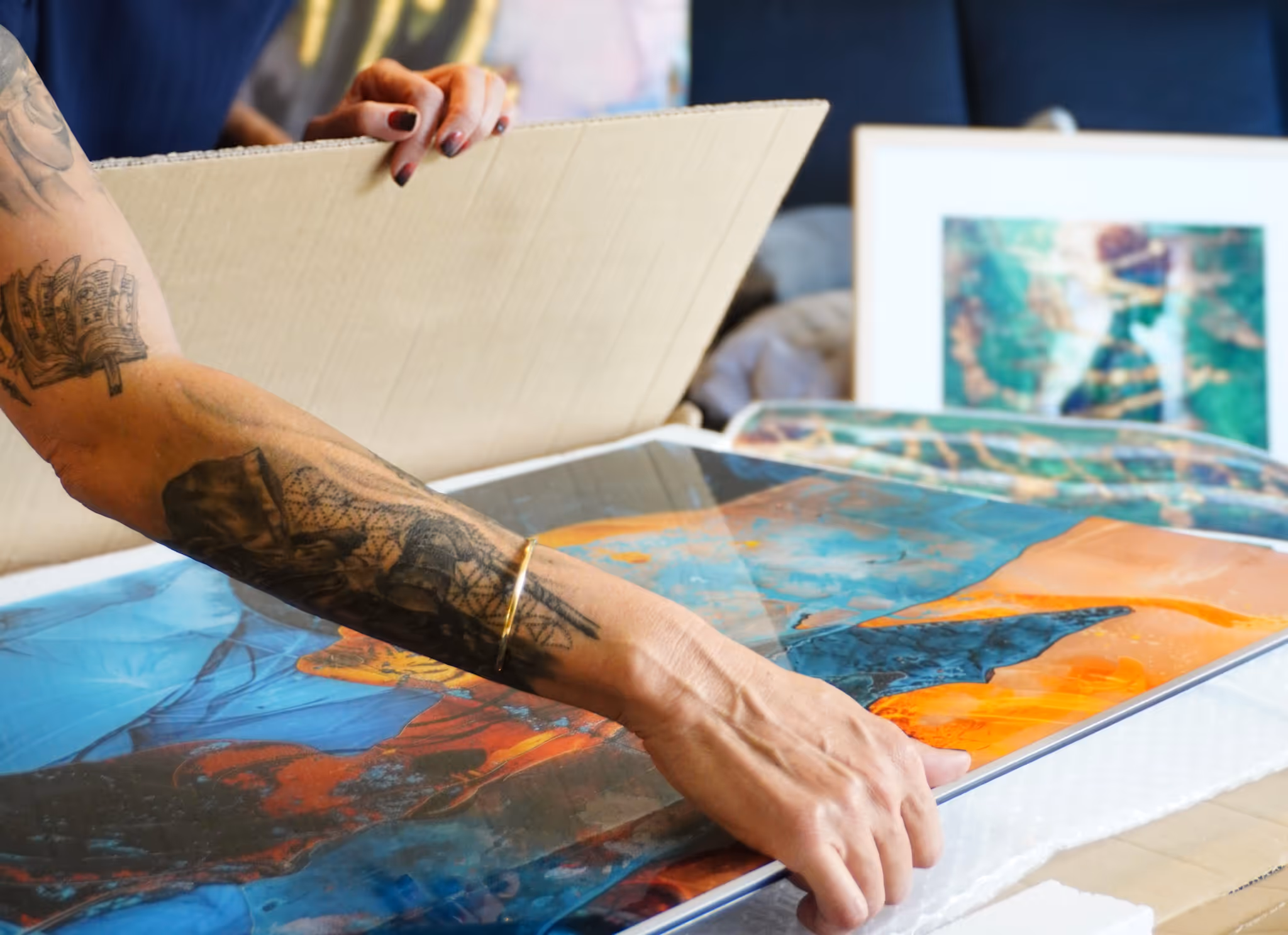 Person mit tätowiertem Arm verpackt ein farbenfrohes Kunstwerk in Karton.