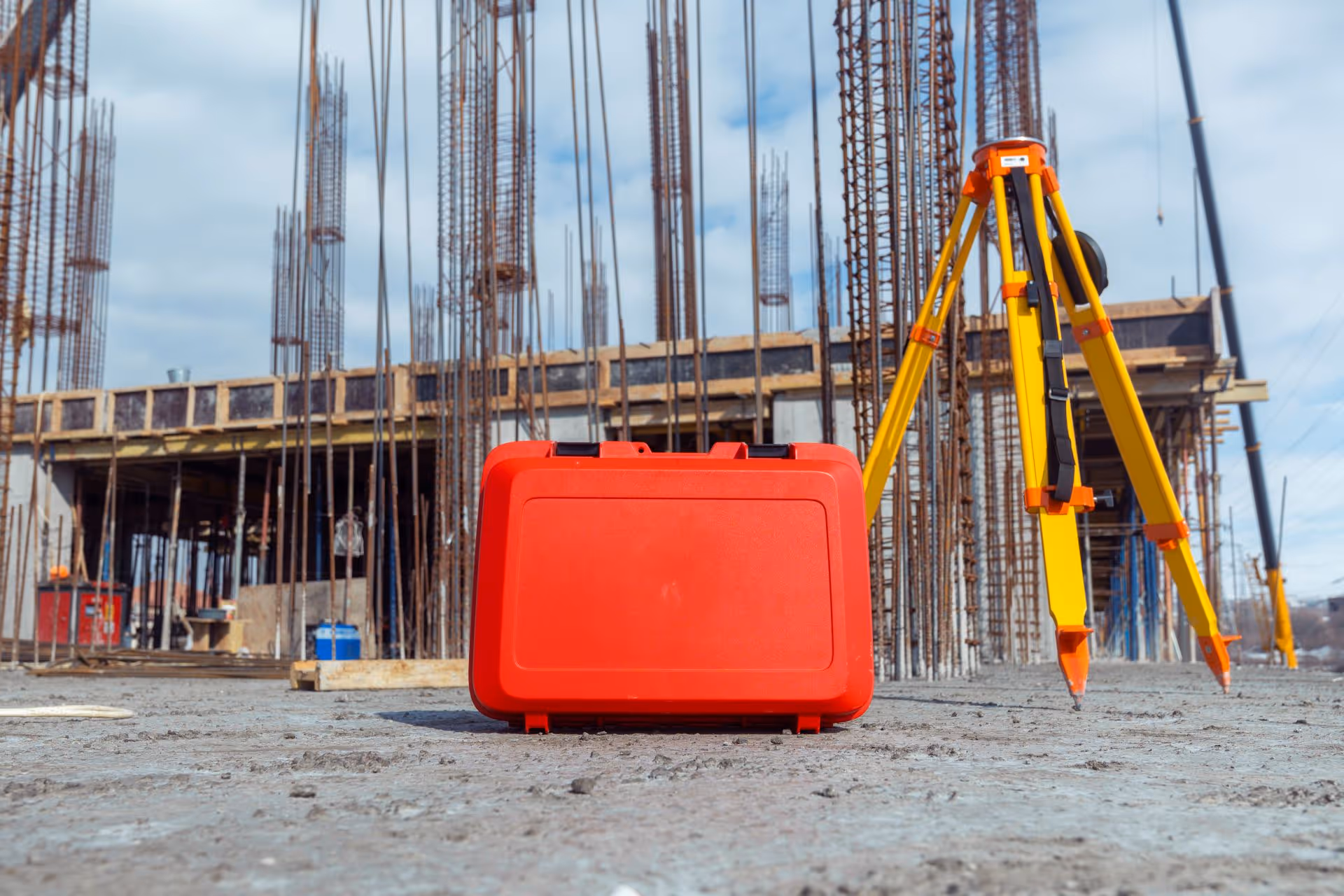 Red equipment case and yellow tripod stand on concrete floor at a construction site with steel rebar columns.