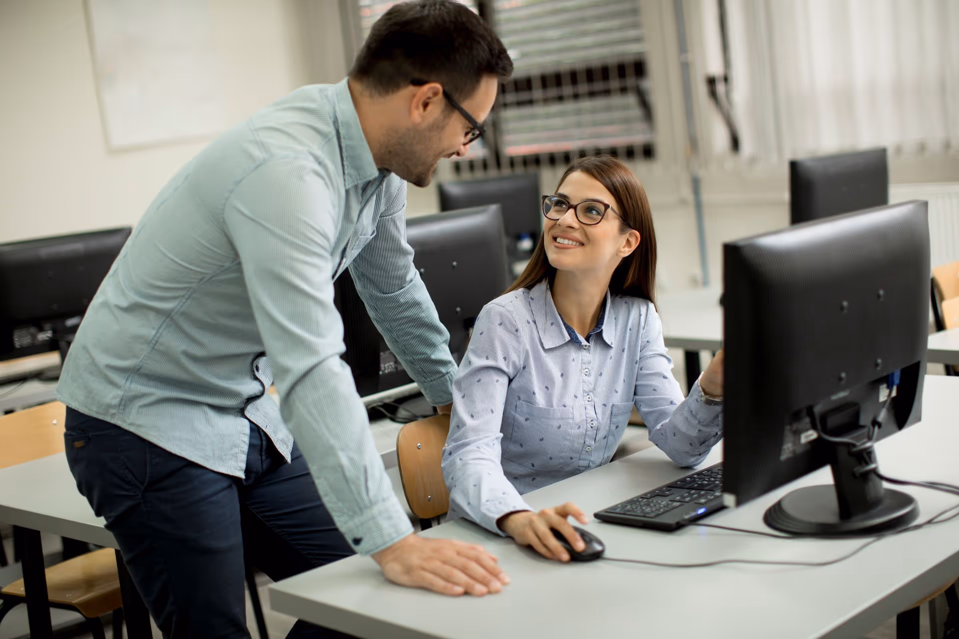 Woman sitting at a computer desk smiling and looking up at a man who is standing beside her in a classroom setting.
