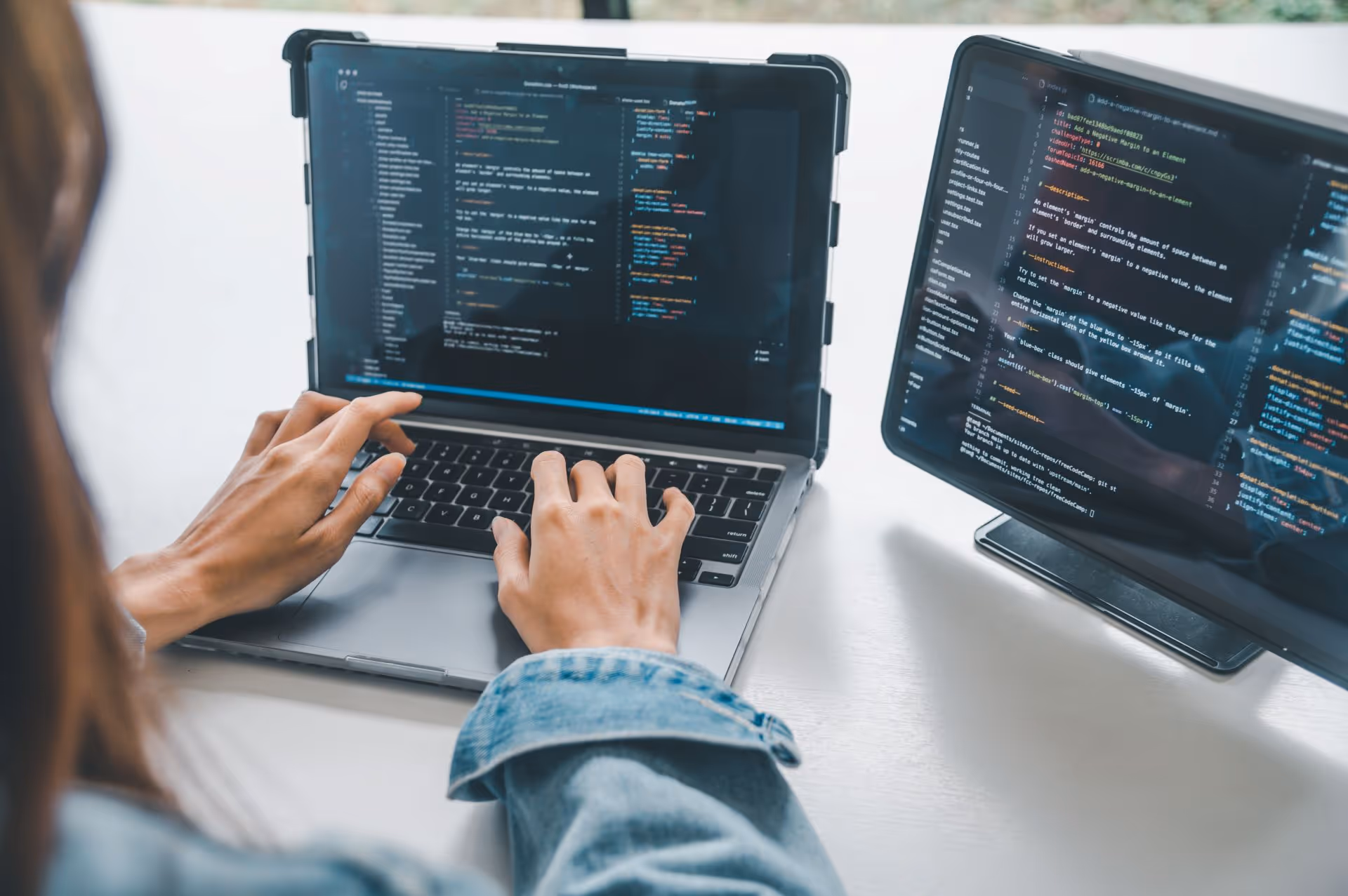 Person typing code on a laptop with a second monitor displaying coding scripts on a white desk.