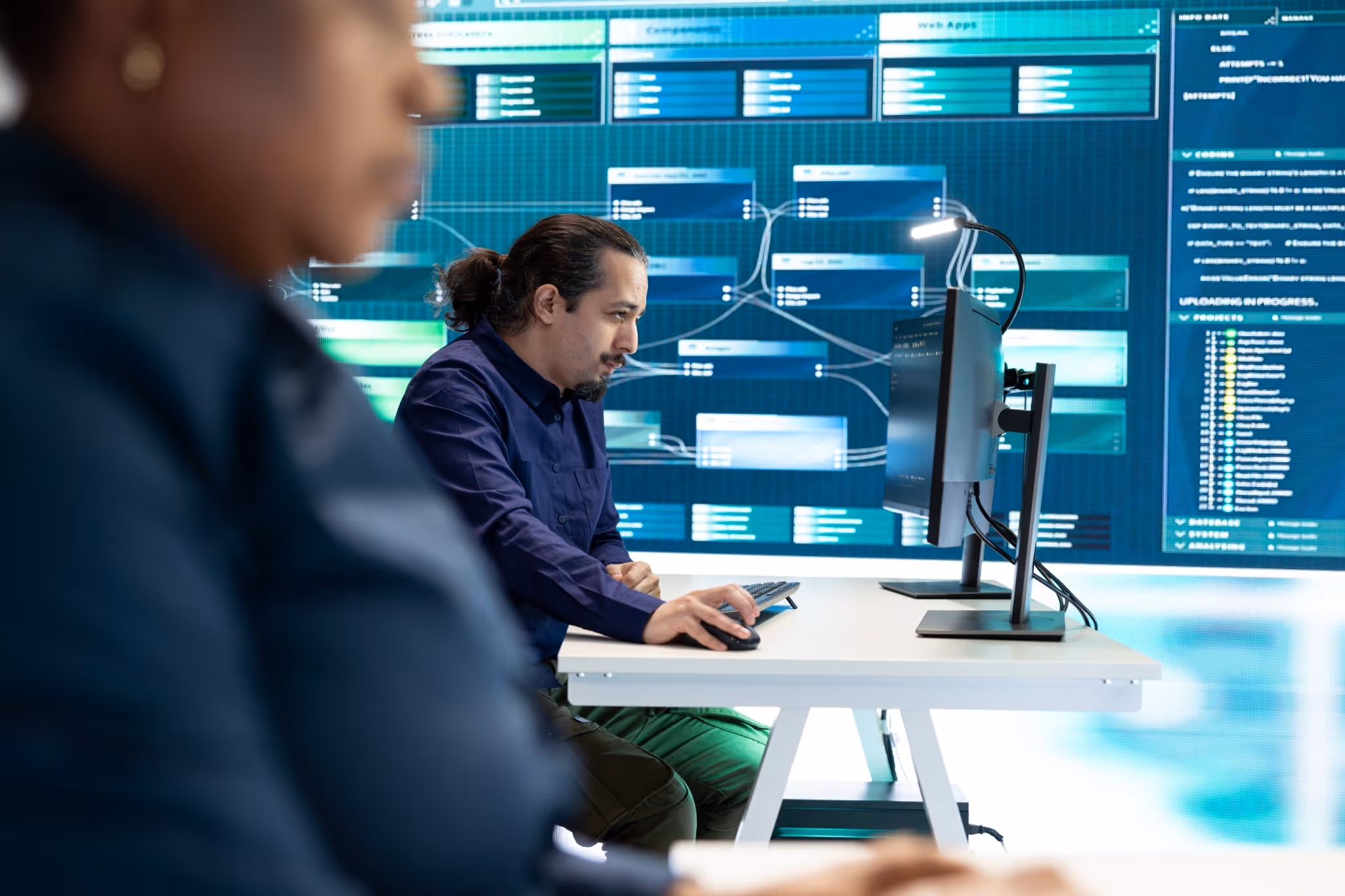 Man with a ponytail working on a computer with technical diagrams and coding visible on large screens behind him.