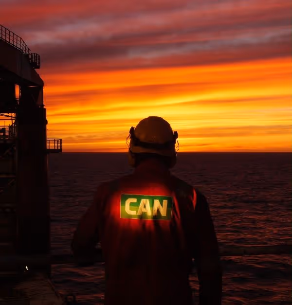 Person wearing a helmet and red jacket with 'CAN' on the back, standing on a platform overlooking the ocean at sunset.