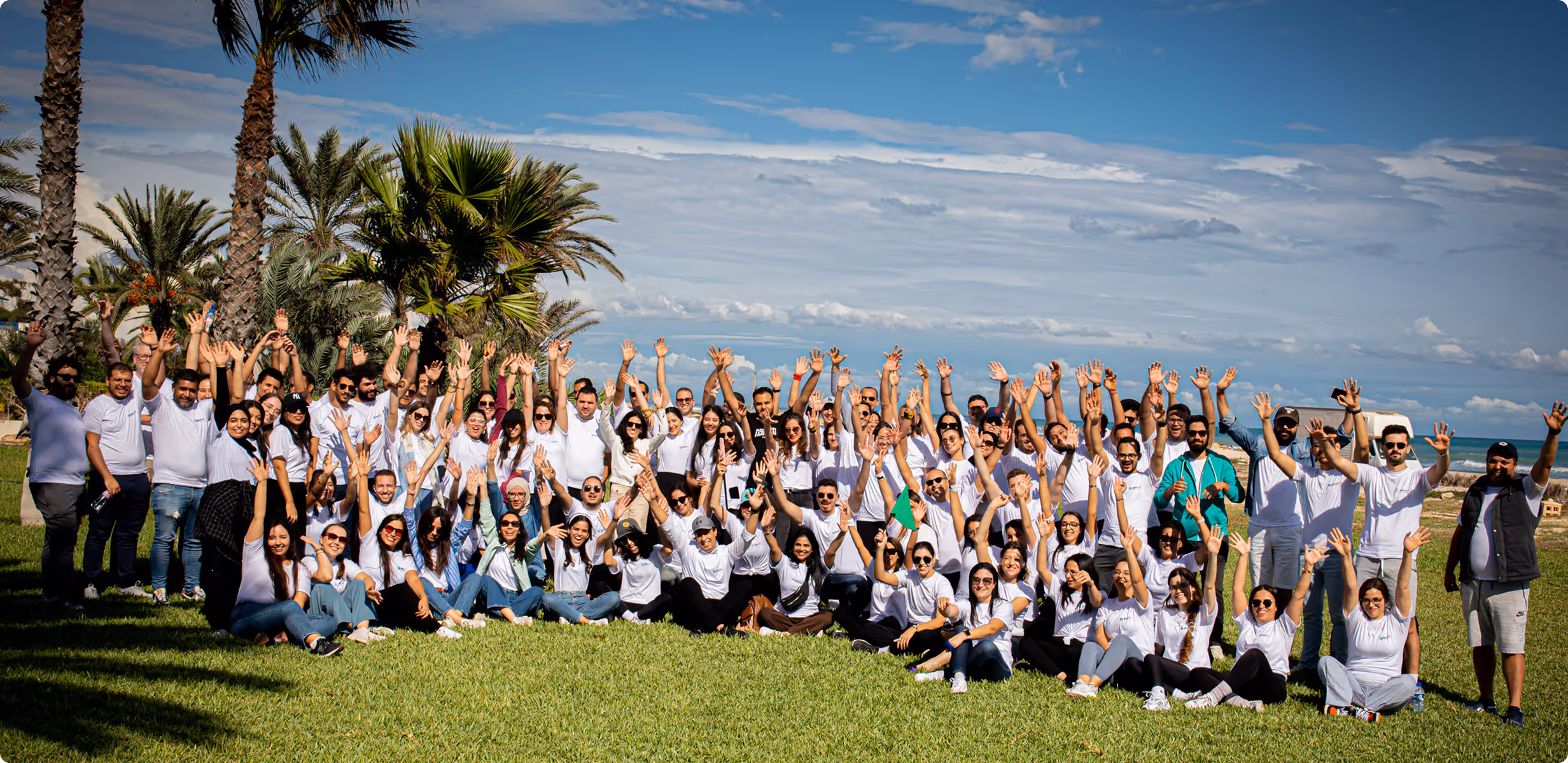 Large group of people outdoors on grass with raised hands under a blue sky and palm trees.